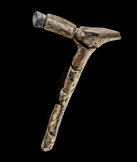 An axe featuring a stone head fitted into an antler sleeve, which is attached to a weathered wooden handle. The tool is displayed against a black background, highlighting its rough, handcrafted construction and aged materials.