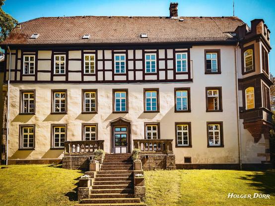 Die Vorderseite des Schlosses Großen-Buseck mit symmetrischer Fachwerkfassade, breiter Freitreppe und gepflegtem Rasen im Sonnenlicht – ein stiller Blick auf gelebte Geschichte.