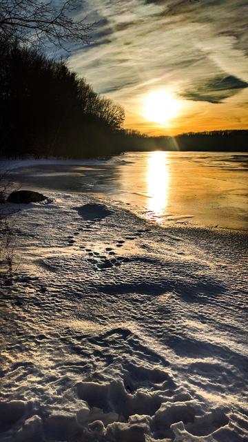 Standing near the shore of a frozen lake. The ground around us is coated with an inch or two of snow that has melted and refroze, capturing small animal tracks and other details in the ice. The sun is setting over the wooded far shoreline, casting deep shadows across our miniature tundra. Leafless trees in the forest surrounding the lake are silhouetted in the light. The sky is filled with streaks of hazy thin clouds, tinted gold and orange along the horizon. The sun is an enlarged blob of bright yellow behind the veil of haze, creating an elongated bright reflection on the ice.