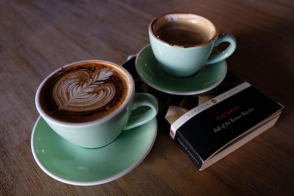 Two coffees on a wooden table. The larger one is in front and has nice latte art. Slightly behind it is a book. On the book is the second coffee which has been partly drunk.