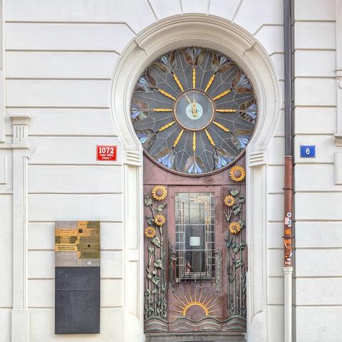 A ground-level view of an Art Nouveau building facade in Prague, showcasing a unique wooden door adorned with carved sunflowers and rising sun motifs. Above the door, a large, round stained-glass window features radiating patterns and a central clock face. The building's white facade has horizontal striations. To the left of the door is a red plaque with white text "1072 STARÉ MĚSTO PRAHA 1" and a metal information board. To the right, a blue plaque displays the number "6," and a copper pipe runs vertically along the wall.