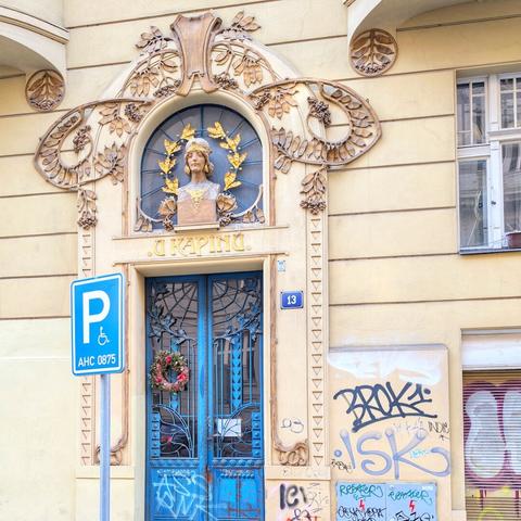 A ground-level view of an Art Nouveau building facade in Prague, showcasing a unique wooden door adorned with carved sunflowers and rising sun motifs. Above the door, a large, round stained-glass window features radiating patterns and a central clock face. The building's white facade has horizontal striations. To the left of the door is a red plaque with white text "1072 STARÉ MĚSTO PRAHA 1" and a metal information board. To the right, a blue plaque displays the number "6," and a copper pipe runs vertically along the wall.
