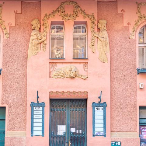 A detailed view of the Art Nouveau facade of Gorazdova 11 in Prague. The building, painted in a soft pink hue, features ornate stucco decorations including stylized figures flanking two arched windows on the upper level and a relief of a reclining lion above the central doorway. The entrance consists of dark teal double doors with decorative metalwork. To the left and right of the doors are teal plaques with white text, one displaying "1995" and the other "№ 11". Swirling decorative elements and floral motifs are integrated throughout the facade.