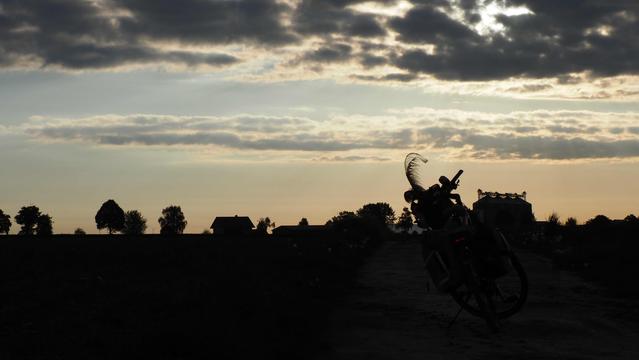 Ein paar Wolken am Morgenhimmel, man erkennt einen Feldweg und ein Fahrrad mit Federn am Lenkerkorb und zwei Packtaschen, als Schattenriss sind silos , Bäume und ein Haus zu erkennen