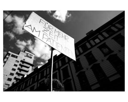 The black and white, low-angle shot focuses on a rectangular sign held aloft on a metal pole. The sign reads 'FÜR MEHR ZEIT FÜR PATIENTEN' in handwritten, capital letters. Below this, 'IM' and then 'AMBULANTEN' are written. The sign is tilted upwards against a dramatic sky filled with dark, textured clouds and patches of brighter areas. In the blurred background, tall buildings with multiple windows are visible, suggesting an urban environment. The person holding the sign is only partially visible at the bottom, showing dark, curly hair. The overall impression is of a protest or demonstration advocating for more time for patients in outpatient care.