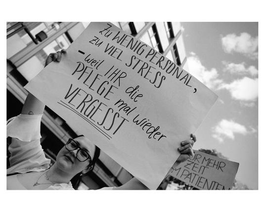A low-angle, black and white photograph captures a person holding up a protest sign. The sign is made of a light-colored material and features handwritten text in dark ink in German. The text is stacked in several lines and reads: "ZU WENIG PERSONAL," followed by "- weil IHR STRESS," then "PFLEGE mal wieder," and finally "VERGISST."

The person holding the sign appears to be young and is wearing glasses and a light-colored top with ruffled sleeves. They are looking upwards towards the sign. Their arms are raised, holding the sign in front of them. A necklace is visible around their neck.

In the background, out of focus, are tall buildings and a cloudy sky. Another partial protest sign is visible on the right side of the frame, with the words "FÜR MEHR ZEIT FÜR PATIENTEN" partially legible. The overall impression is of a demonstration or protest taking place in an urban environment.