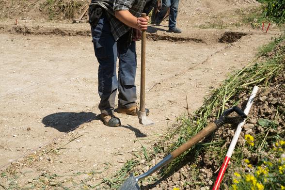 Alt text : the camera is focused on the legs of one of the archaeology team, they were banging the pick on the floor, presumably to get excess dirt off before going back to work. His boots and trousers are dusted with the same dirt that can be seen all around, the trench behind them bright with chalk.