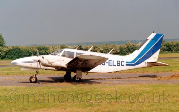 Side view of a white, twin propellor-engined light aircraft parked on tarmac facing to the left.
The plane is mostly white, with a pair of thin stripes, one light blue, the other dark blue, running along the body, getting wider as the move rearwards, until they sweep up to fill most of the tail.
The dark blue registration is on the rear fuselage, as well as under the left wing.
Green grass fills the foreground, with trees stretching across the background, under grey skies.