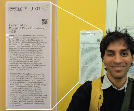 Vidur Mithal in front of a sign saying "Hasselmann Hall U-01/Konferenzsaal" and a short description of Hasselmann's contributions to climate science.