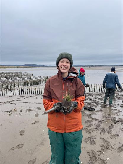 smiling person holding a plant, standing on an area of mud with saltmarsh planting taking place. two people are in the background, there are fences in the mud to protect the plants as they establish.