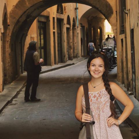 Teen brunette model Emily posing under arch way on street in Florence Italy in a light floral dress with hand on hip. Long dark hair is braided. People in dark street in background.