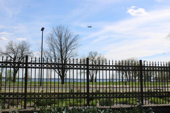 Wrought iron fence with flower beds on both sides before a paved road with a tall street light with a green lawn, starting to bud trees next to a blue lake under partly cloudy sky with an airplane in the process of descent.