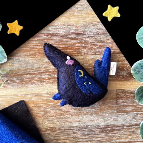 A little handmade felt plushie photographed on a black and wooden background, surrounded with decorative plants, and felt stars.  The plushie is of a black raven with dark blue wings, tail and feet. It is looking up with a white embroidered eye, and a soft pink blush. Three stars and a crescent moon are embroidered on its wing.