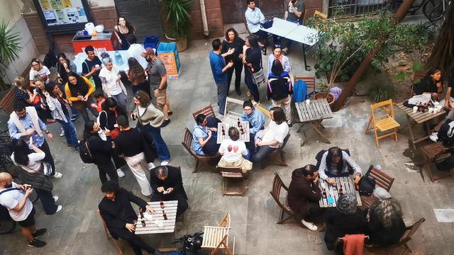 Vista del patio con participantes en el evento. Algunos sentades en mesas otres de pie. La foto está hecha deste el primer piso.