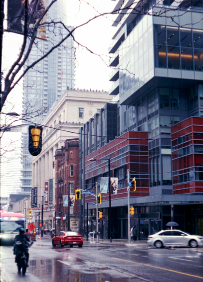 A colour photograph of a grey day on a downtown street - new developments squeeze the old as roadwork and construction squeezes cars, buses, and bikes.