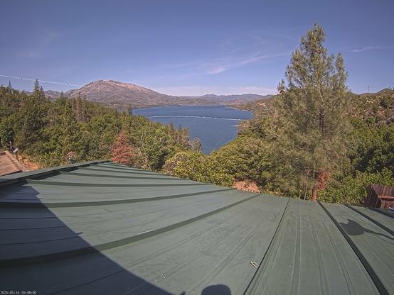 Whiskeytown Lake in Whiskeytown National Recreation Area. The roof of the visitor center appears in the foreground. Trees and shrubs fill the center of the image. The lake sits dead center, connecting the greens and reds of the foreground to the tans and greys of the mountains in the distance. The sky today is mostly blue. Some clouds can be seen, especially on the far horizon.