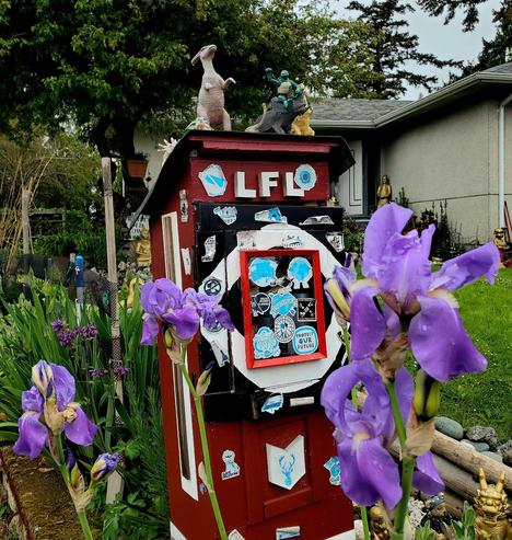 Eye-level, medium shot capturing a roadside art installation. A red and black painted wooden structure, reminiscent of a small building, dominates the center. It is decorated with stickers, with text and images. The structure is topped with toy figurines. In the foreground, out-of-focus purple irises with water droplets soften the composition and guide the eye towards the installation. The background includes garden vegetation and a residential house, but it remains blurred to maintain focus on the artwork.