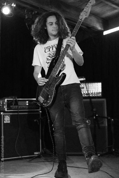 A musician with curly hair plays a bass guitar on stage, wearing a graphic t-shirt and denim jeans. The background features music equipment, with focused stage lighting creating a dynamic atmosphere.