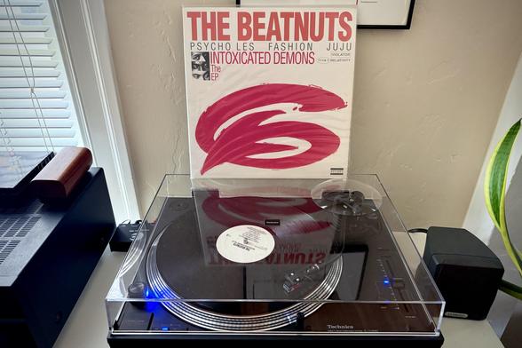 A black vinyl record spins on a black turntable. The record sleeve stands up on turntable’s closed dust cover behind the platter.