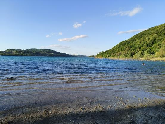 Photographie numérique couleur en format paysage.
Un lac dans des tons de bleu et de brun, agité de vaguelettes, avec en face et à droite des collines boisées. Le ciel est bleu avec quelques nuages blancs, avec une lumière de fin d'après-midi.