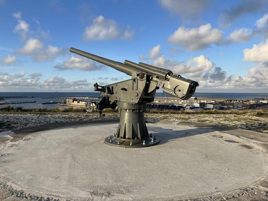 The image shows a large, mounted artillery gun situated on a concrete platform, likely part of a historic coastal defense installation. In the background, you can see a body of water, possibly a sea or large lake, and what appears to be a port area with various buildings and structures. The sky is partly cloudy, suggesting a bright, clear day which enhances the visibility of the surroundings. The presence of this gun suggests the location has military historical significance.