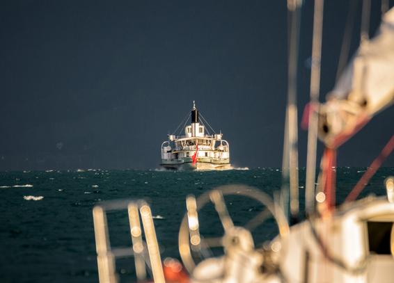 Im Vordergrund ist unscharf ein Teil eines grossen Segelbootes abgebildet, im Hintergrund entfernt sich ein Schaufelraddampfer auf einem stürmischen See.