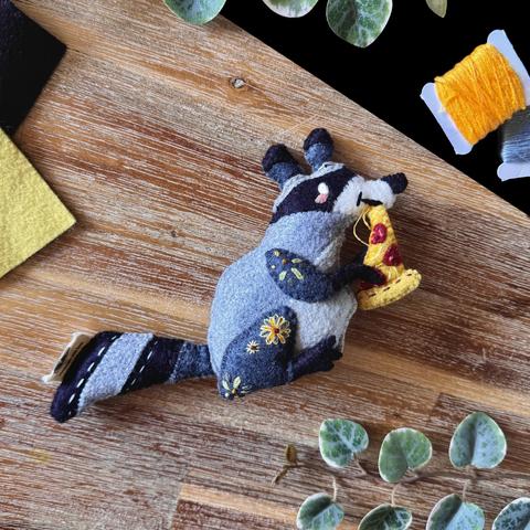 Picture of a felt raccoon plushie photographed on a black and wooden background, surrounded with decorative plants and matching threads and felt. 
The raccoon is munching on a slice of pepperoni pizza, threads are emulating the string of cheese between the pizza and its mouth. 
Yellow flowers and green leaves are embroidered on its legs.