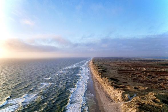This image features a beautiful aerial view of a coastline. On the left, the ocean displays white waves breaking gently onto a sandy shore. The coastline stretches into the distance, meeting a horizon where the sky is soft with the hues of early morning or late afternoon sunlight. To the right of the beach, the landscape showcases dunes and rugged terrain with sparse vegetation, which then extends into a flat, expansive landscape further inland. The contrasts between the blue water, sandy shores, and the terrain create a serene and natural scenic view.