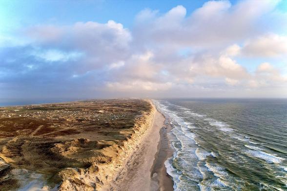 This image features an aerial view of a coastal landscape. The scene shows a sandy beach edging a vast ocean, with waves gently breaking along the shoreline. The left side of the image displays a diverse terrain covered with dunes and sparse vegetation, indicating a somewhat arid environment. The sky above is partly cloudy, casting soft lighting over the area, with hints of blue sky breaking through. The beauty of a long stretch of coastline and the natural contrast between land and sea are showcased prominently in this photograph.