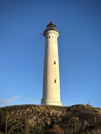 The image features a tall, white lighthouse situated on a grassy hill. The lighthouse has a classic cylindrical shape with a red dome on top, and a small antennae structure attached to one side. It's a clear day with a blue sky in the background. This setting suggests a coastal location, as lighthouses are typically used to aid in naval navigation.