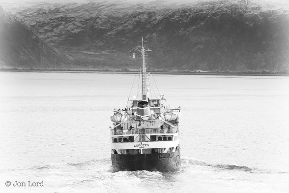 This Is A Black And White Seascape / Maritime Photo In Landscape Format. Rising Upward To The Two-Third Point Of The Frame Is A Body Of Calm Water. Below The Centre Of The Image Is A Stern View Of A Medium Sized Sea Going Ferry. The View Is Aligned With The Ship's Axis, So The Stern And Topsides Are In View: The Sides Are Not. Like So Many Merchant Ships The Hull Is Black While The Superstructure Is White. The Stern Is Rounded As Is The Superstructure Immediately Above. The Ships Name 'MS LOFOTEN'  Is In Bold Black Letters Above The Hull Together With Her Port Of Registry 'Tromsø'. Above The Black Hull Are Three Decks. A Few Passengers Can Be Seen On The Upper Two. On That Upper Deck There Are Four Open Lifeboats Hanging From Davits, Two On Either Side. There Is A Single, Small Raked Funnel Aft But Without Any Smoke Or Fumes Visible. Forward Of The Funnel Is A Tall White Mast With A Number Of Aerials And The Shipping Line's Pennant. Ahead Of Our Ship And Perhaps Two km Away Is A Steep And Rocky Cliff, The Upper Part Covered In A Light Dusting Of Snow. 
Our Ship Was In Service With The Hurtigruten Coastal Express  Between Bergen And Kirkenes From 1964 To 2020 And Has Now Been Repurposed As A Training Ship. The Location Is In The Breisundet, 71º North Of The Equator, High In Norway's Arctic North (2018).