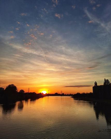 A serene sunset over a calm river. The sun, a brilliant orange orb, is positioned low on the horizon, casting a vibrant golden and orange glow across the water's surface. The sky is a blend of colors, transitioning from a deep blue at the top to lighter hues of blue and streaks of pink and orange closer to the horizon. Wispy clouds are scattered across the sky. Silhouetted trees and buildings line the banks of the water.