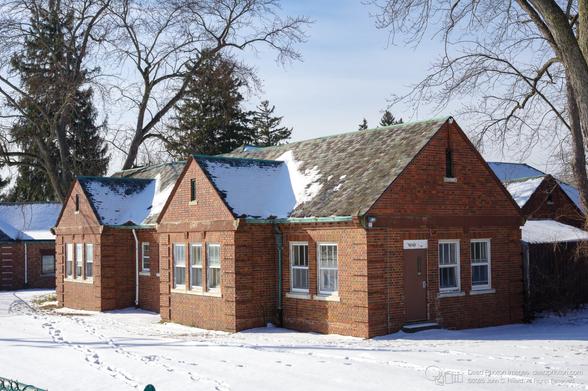 The image shows a low, red brick building with a complex roofline, partially covered in snow. The building appears to be composed of several connected sections, each with a gabled roof. The roof is made of multi-colored slate tiles, with patches of snow clinging to the surface. The brickwork is a deep red, with some variation in color and texture. Windows are visible on the front of the building, with white frames. The ground is covered in a thick layer of snow, with visible footprints leading towards the building. Bare trees frame the building against a pale blue sky. The overall impression is one of a sturdy, somewhat aged structure in a winter landscape. The building's architecture suggests a historical or institutional purpose, possibly a school or community center. The presence of snow and bare trees evokes a sense of quiet and stillness.