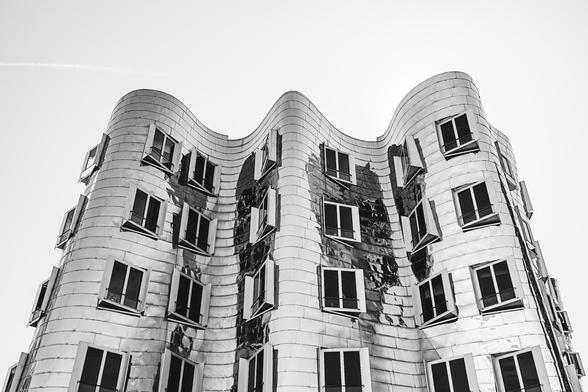 Black and white image of a curving, futuristic-looking building clad in reflective metal panels, with square windows partially opened. The architecture features a fluid, wave-like facade, creating a sense of motion and distortion.