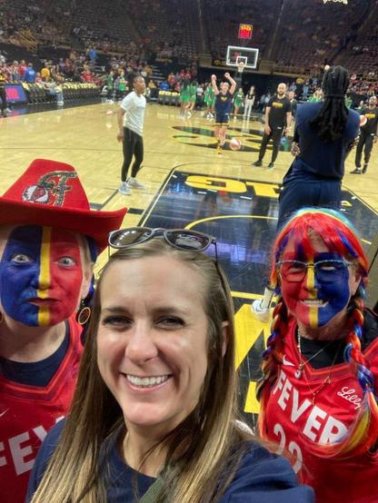 Image is a selfie of two insane Indiana Fever fans with ESPN courtside  reporter Holly Rowe, the fans done up in red, blue and gold facepaint and Fever jerseys, the one on the left wearing a red Fever cowboy hat and making a very dorktacular version of a "gameface."