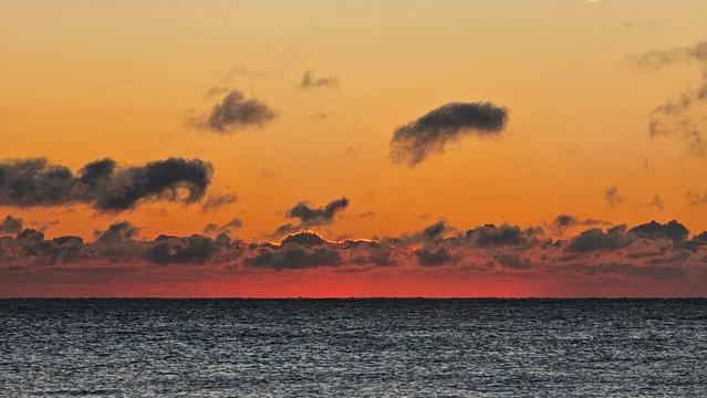 A wide shot of the ocean at sunrise. The horizon line shows a dark, rippling sea meeting a vibrant orange and yellow sky with scattered dark clouds. The sun is just beginning to appear above the horizon, casting a bright red glow across the water and the undersides of the clouds.