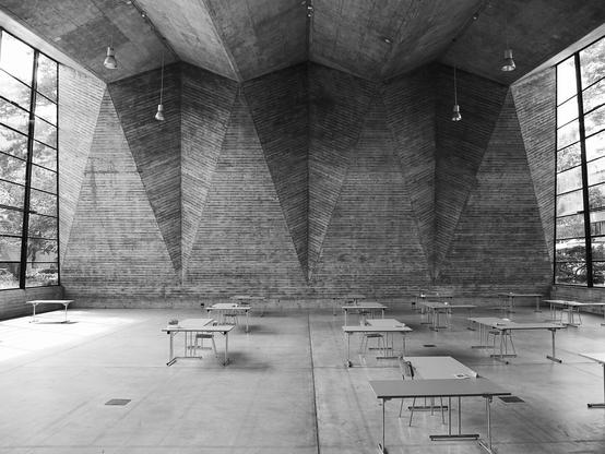 A minimalist interior space with a dramatic geometric concrete ceiling featuring angular facets and pendant lights. Floor-to-ceiling windows along both sides allow natural light to illuminate the sparse room containing several simple tables arranged on a concrete floor. The stark brutalist architecture creates a sculptural quality to the space. (made with help of claude.ai)