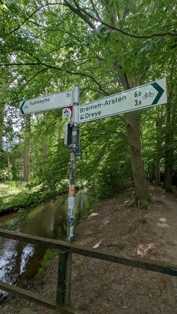 Foto im Wald an einer Brücke, im Vordergrund das Brückengeländer, daran ein Schild. Nach links: "Sudweyhe, 0,9km", nach rechts: "Bremen-Arsten 6km und Dreye 3,0km" daneben jeweils ein Fahrradsymbol. Die Brücke geht über einen Bach, der gesamte Hintergrund besteht aus grünen Bäumen