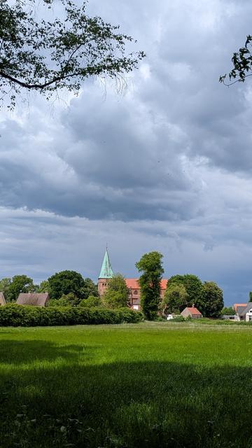 Blick über ein grünes Feld auf eine ca. 100m entfernte Kirche, links und rechts daneben sind die Dächer von Wohnhäusern zu sehen, dunkelblauer, bewölkter Himmel, links und rechts oben Äste von Bäumen