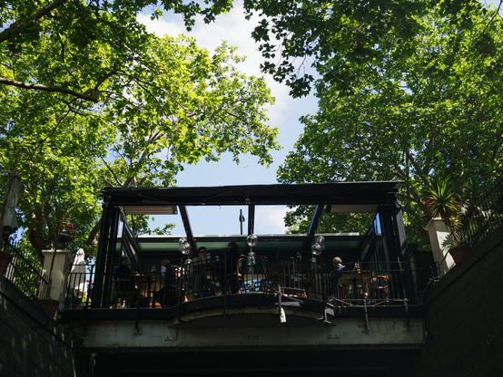 An outdoor café sits on a platform over a city channel surrounded by lush green trees, with people dining under a sunny sky.