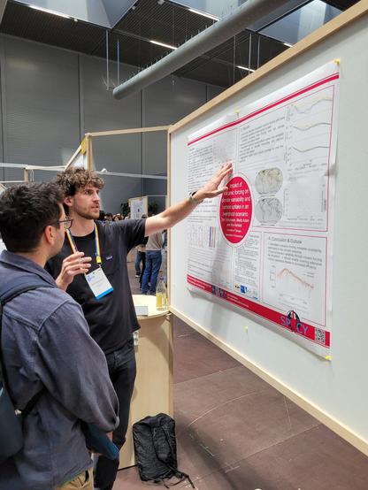 Tom Schürmann zeigt auf ein Poster auf dem die Ergebnisse seiner Forschungsarbeit zu sehen sind und erklärt diese einem Konferenzteilnehmer.