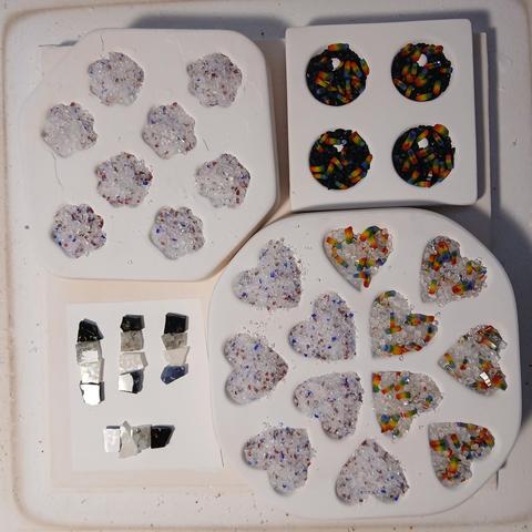 a photo taken from above of three varieties of molds containing small fragments of crushed glass: scalloped, circular pendant, and heart-shaped. The clear crushed glass is dotted with pink and blue pieces in the scalloped and half of the heart shapes, with small rainbow-striped rods in the other half of the hearts, and the same rainbow rods are mixed with a dark blue-black glass in the pendant shape.

There is also a square in the lower left of four groups of four square glass pieces in lines, in gradients of dark to light.