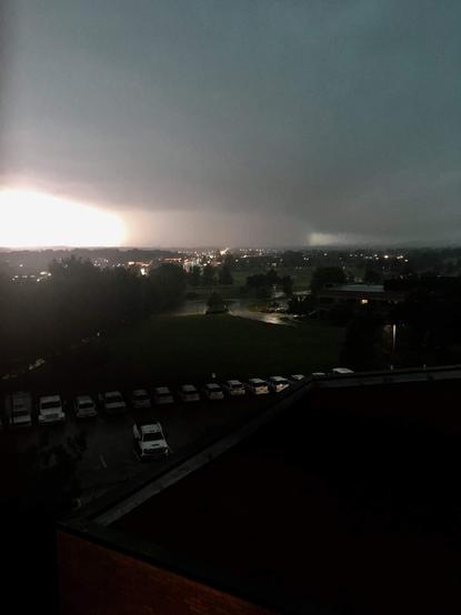A photograph of the May 2019 tornado south of town. This was taken from campus, which is on a hill. The city spread out south toward the tornado is unnaturally dark at 7pm because of the rain clouds, and the sky is obscured by clouds full of rain, but in the distance, you can see a huge, rain-wrapped funnel on the ground which is backlit by a bright southern sky.