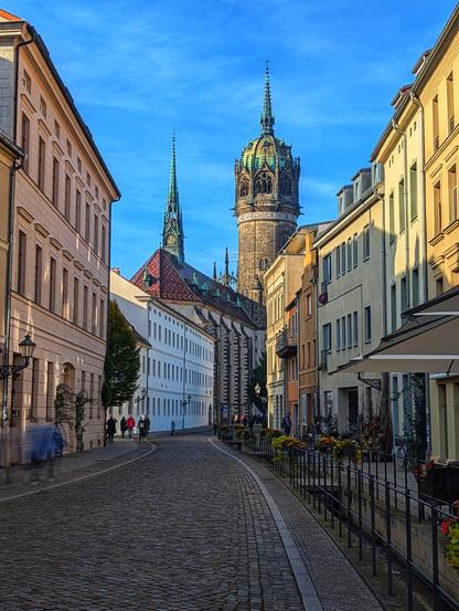 A cobblestone street curves gently between buildings of varying heights and colours under a bright blue sky. In the distance, the distinctive towers of the Castle Church in Wittenberg rise prominently. People stroll along the pavements. The scene is bathed in soft daylight, casting subtle shadows on the buildings.