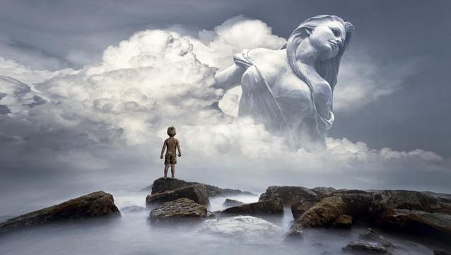 A small boy standing on rocks in the mist, looking at a giant mythical figure of a woman in the clouds.