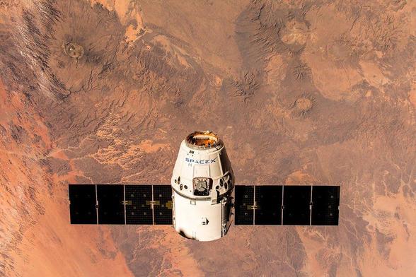 Ein Raumfahrzeug mit der Aufschrift "SPACEX" schwebt im All über einer felsigen, roten Landschaft, die von Kratern und Erhebungen durchzogen ist. Das Raumschiff hat Solarzellenflügel auf beiden Seiten und ist von einer wolkenlosen Umgebung umgeben.
