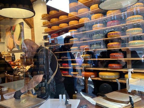 Photograph by Fenichel

A look through a store window on a busy street. It is a cheese store, with the back wall of shelves with many rows of colorful cheese wheels. On the left is a man in black, with short sleeves, black, gloves, slicing cheese. In the center and right are the cheeses in very sharp focus, along with reflections of distant buildings and people walking on the sidewalk or streets.