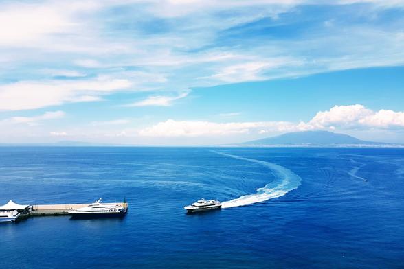 An aerial view of a deep blue ocean with two boats. One boat is docked at a pier on the left, and another boat is moving towards the right, leaving a curved white wake behind it. In the background, a large mountain rises under a partly cloudy blue sky.