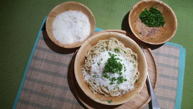 A large banana leaf bowl with two smaller ones diagonally above it. They stand on a wicker placemat. The large one (on a bamboo plate) contains spaghetti topped with pardano and chopped curly parsley. The two smaller bowls contain pardano and parsley. A part of a fork is visible. Tablecloth is green.