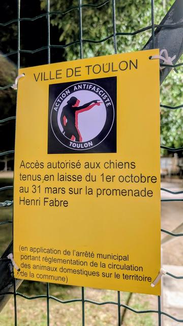 Un panneau jaune accroché à du grillage. Ça dit "Ville de Toulon, accès autorisés aux chiens tenus en laisse du 1er octobre au 31 mars".

Sous le texte "Ville de Toulon" un sticker action antifasciste anarchiste Toulon (avec une statue, le génie de la navigation, qui pointe son doigt vers la droite) à été collé sur le blason de la ville.